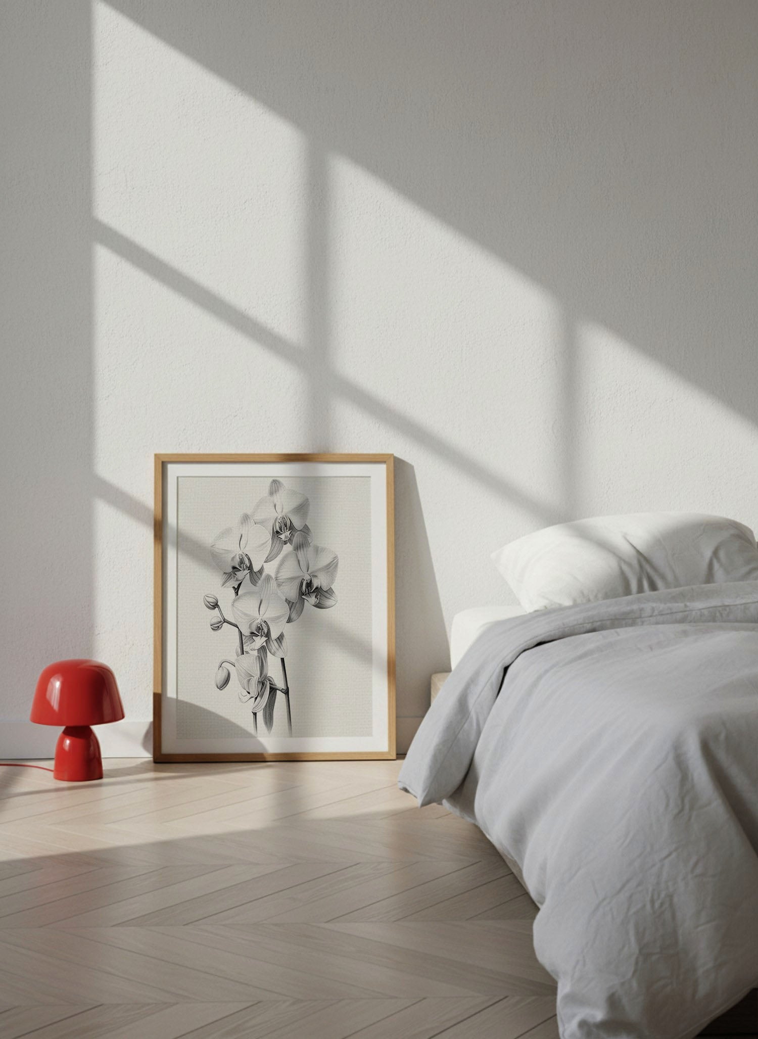 A framed black and white floral art print with a light wood frame leans against a white wall in a minimalist bedroom. Sunlight casts windowpane shadows across the wall and bed, with a small red mushroom-shaped side table on the floor.