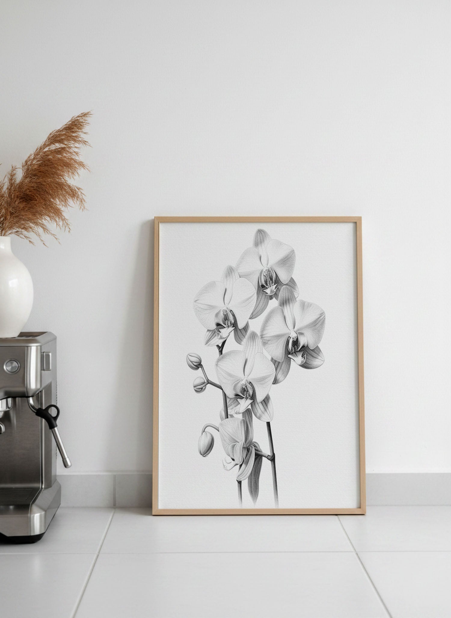 A framed black and white botanical art print of orchids leaning against a white wall on a light tiled floor, next to a silver espresso machine and a white vase with dried grass.