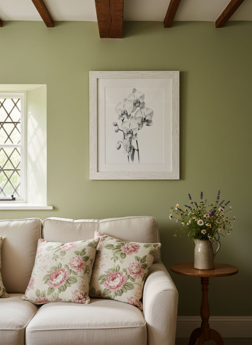 A framed black and white floral art print hangs on a sage green wall above a cream-colored sofa with pink floral throw pillows. A small wooden side table next to the sofa holds a ceramic pitcher filled with wildflowers.