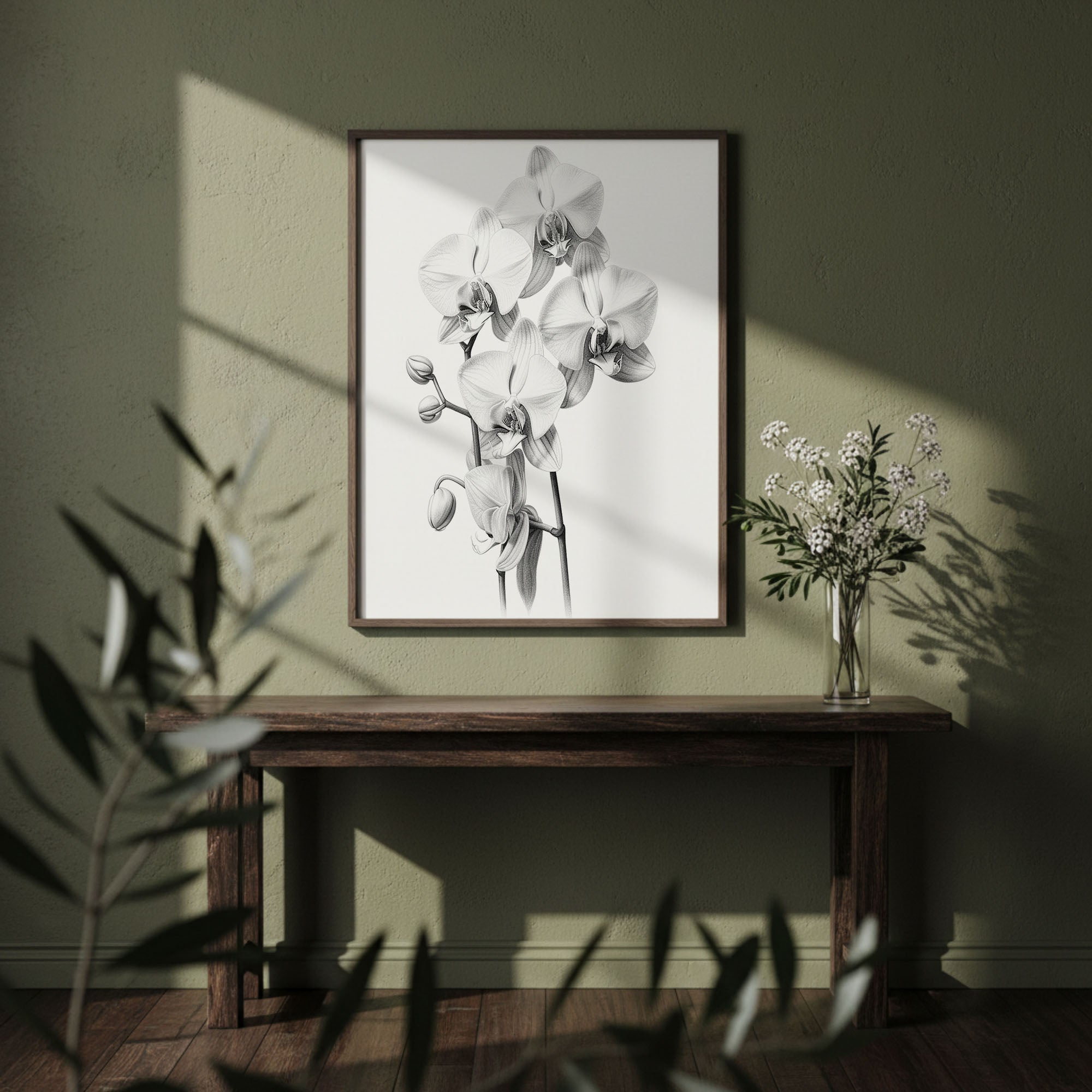 A framed black and white orchid print hangs on a textured olive green wall above a dark wooden console table, styled with a glass vase of white flowers and soft window shadows.
